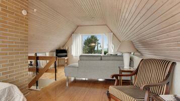 Une chambre confortable située sous les toits, avec un plafond en pente en bois, un canapé gris, un fauteuil à rayures et un téléviseur posé sur une table en bois.