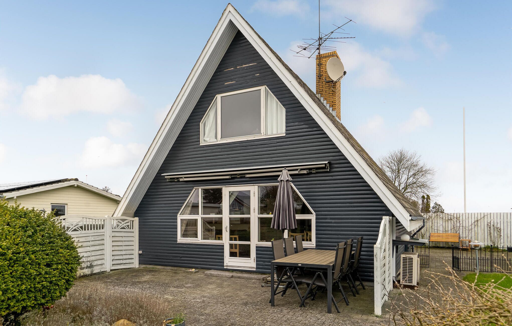 Une maison à deux étages, à l’extérieur sombre, avec des éléments décoratifs blancs et une cheminée. On y trouve une terrasse avec une table et des chaises.