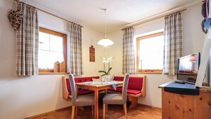 Un rincón comedor acogedor con una mesa de madera, dos sillas y un banco con cojines rojos. Hay una mesita con velas y un jarrón de flores.