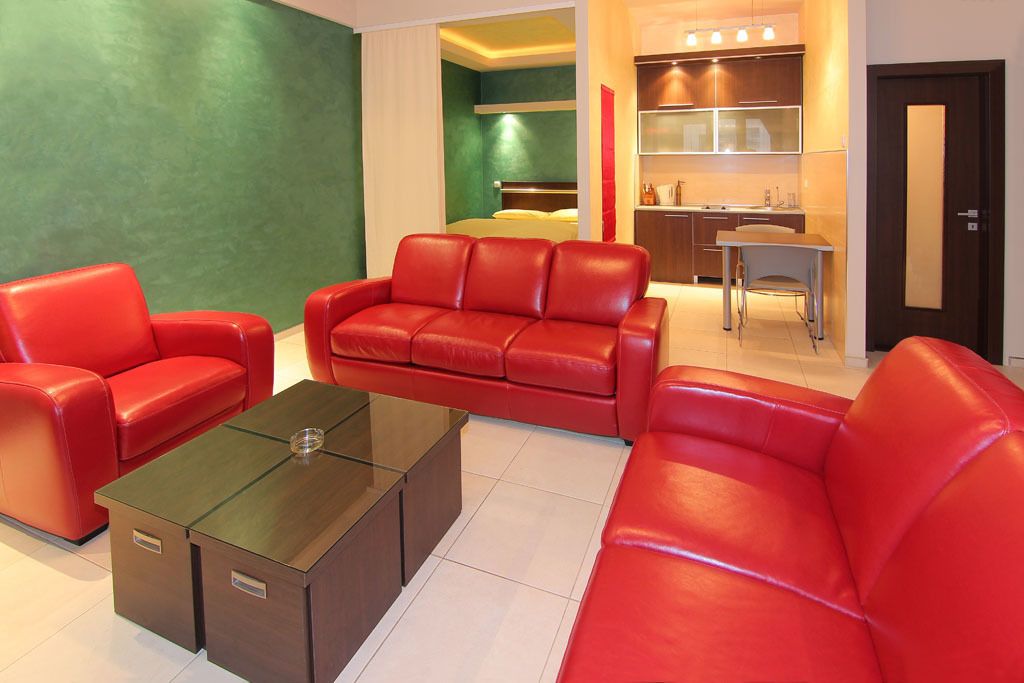 A modern hotel room with red leather sofas, a glass coffee table, a kitchenette, and a green accent wall.