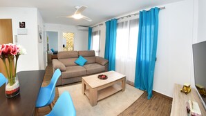 A modern living room with a sofa, coffee table, and blue chairs. There is a TV on the right and a dining area with a table and chairs. - Zenbeach 1A playa Carihuela (Torremolinos)