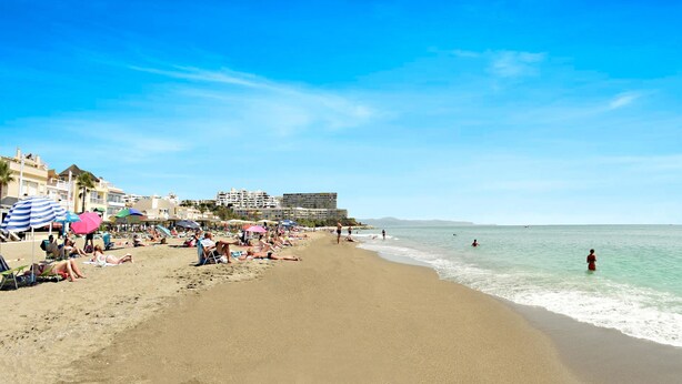 A beach with people sunbathing and swimming, umbrellas, and buildings in the background. - Zenbeach 1A playa Carihuela (Torremolinos)