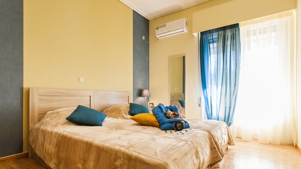 A bedroom with a large bed, a bedside table, an air conditioning unit, and a window with curtains. - Central cozy apartment (Athens)