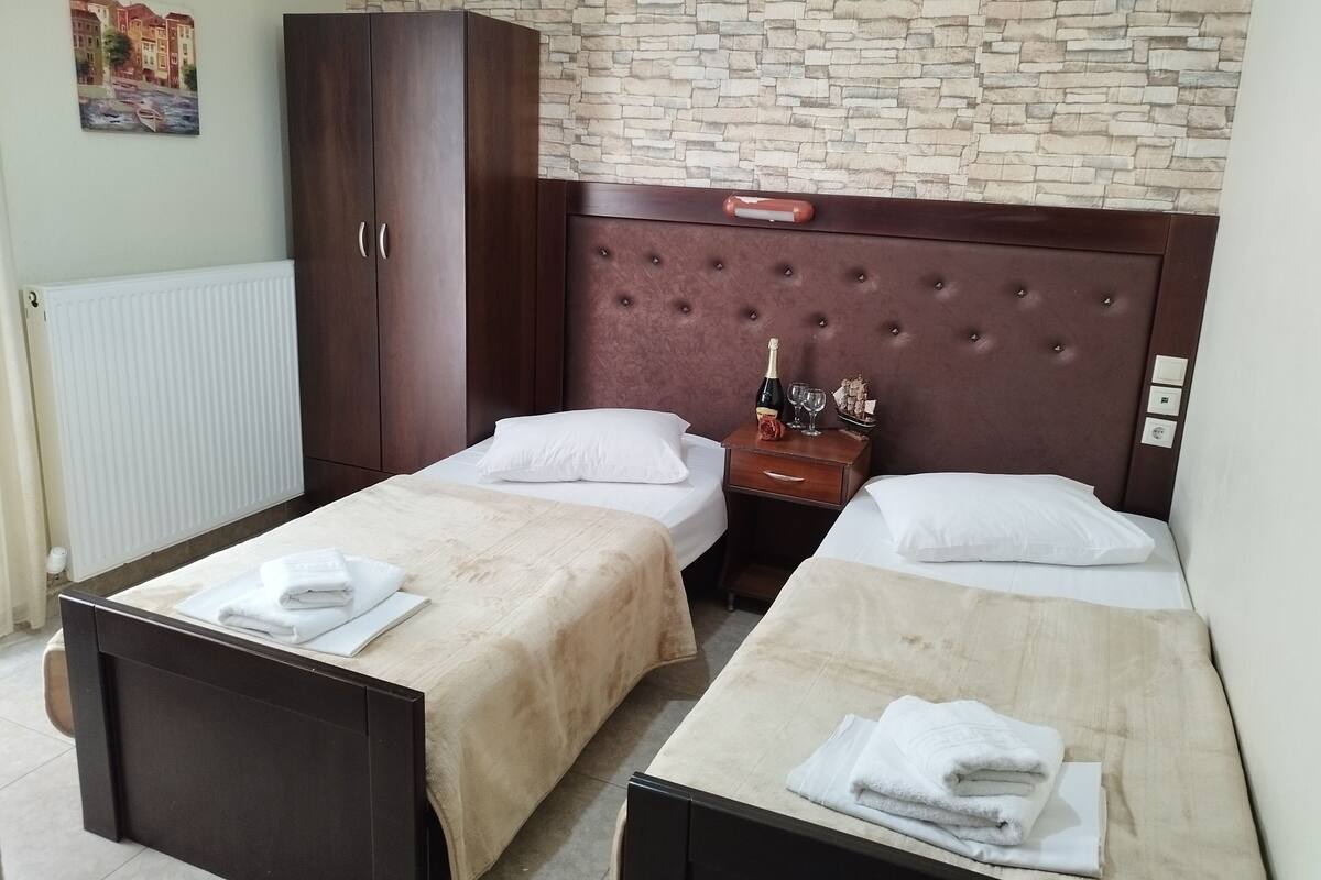 A hotel room with two single beds, a wooden wardrobe, a nightstand with a bottle of champagne, and a stone wall.