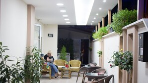 Un área de estar interior con sillas y mesas de mimbre, plantas en macetas y una entrada por puerta de cristal.