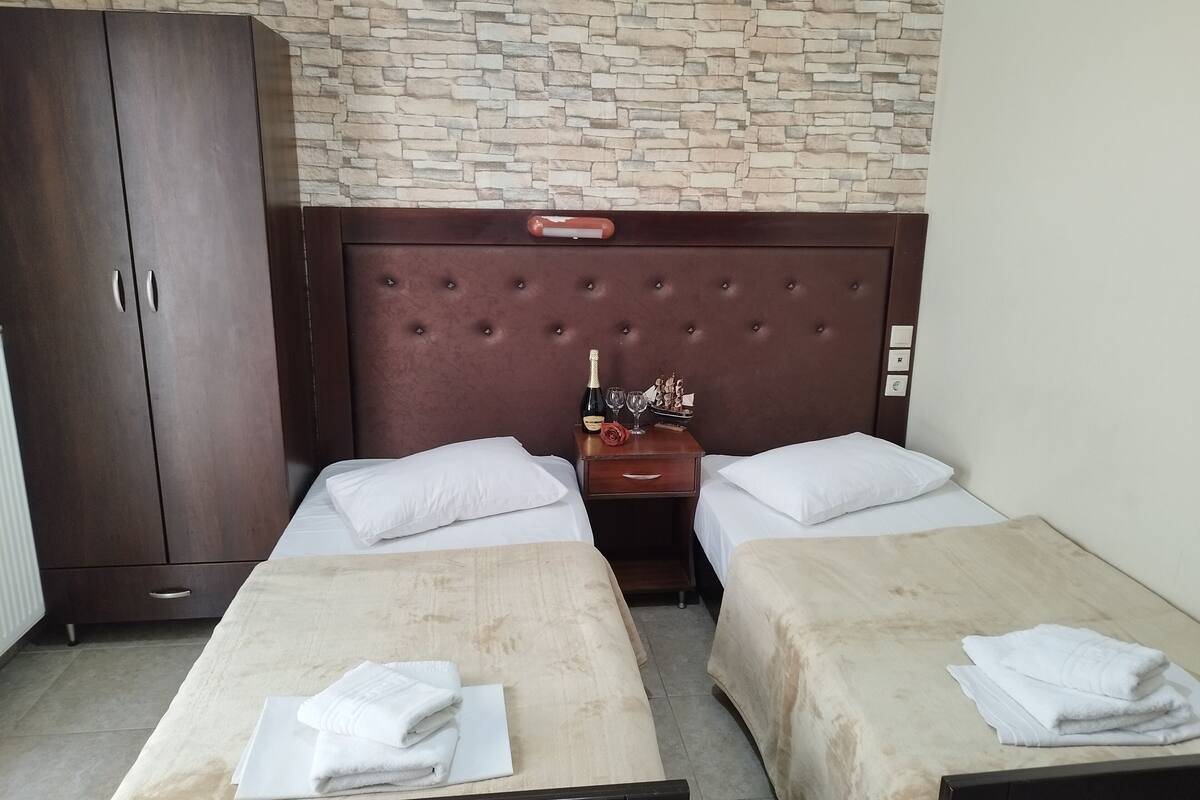 A hotel room with two single beds, a wooden wardrobe, a nightstand with a bottle and glasses, and a stone-patterned wall.