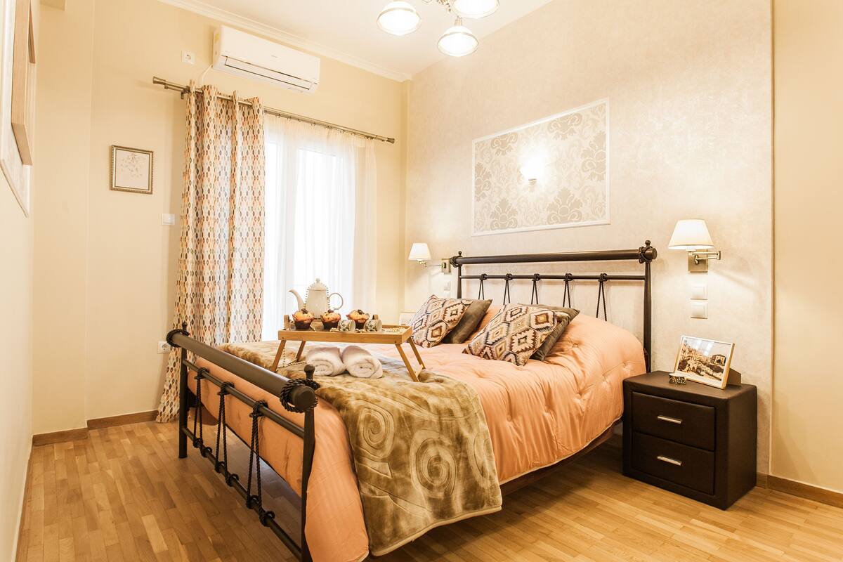 A bedroom with a bed, bedside tables, a nightstand, a window with curtains, and an air conditioning unit.