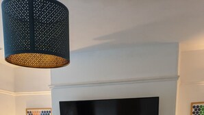 En stue med pejs, fjernsyn ophængt over, sofabord i træ, og et hyggeligt siddeområde med lænestol og sofa.