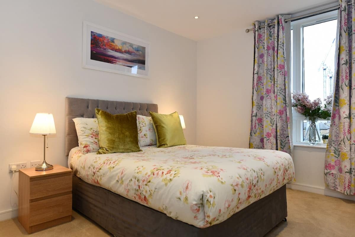 A bedroom with a bed, a nightstand, a lamp, a framed picture on the wall, and a window with curtains.