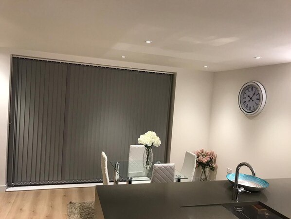 Un espace repas moderne avec une grande table, des chaises, une horloge et une fenêtre équipée de stores.