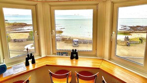 A room with a large bay window overlooking a beach, a wooden desk, and two chairs. - Seafront Apartments (North Shields)