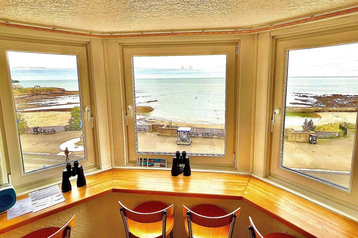A room with a large bay window overlooking a beach, a wooden desk, and two chairs.