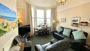 A living room with a large window, a black leather sofa, a dining table, and a chandelier. - Seafront Apartments (North Shields)