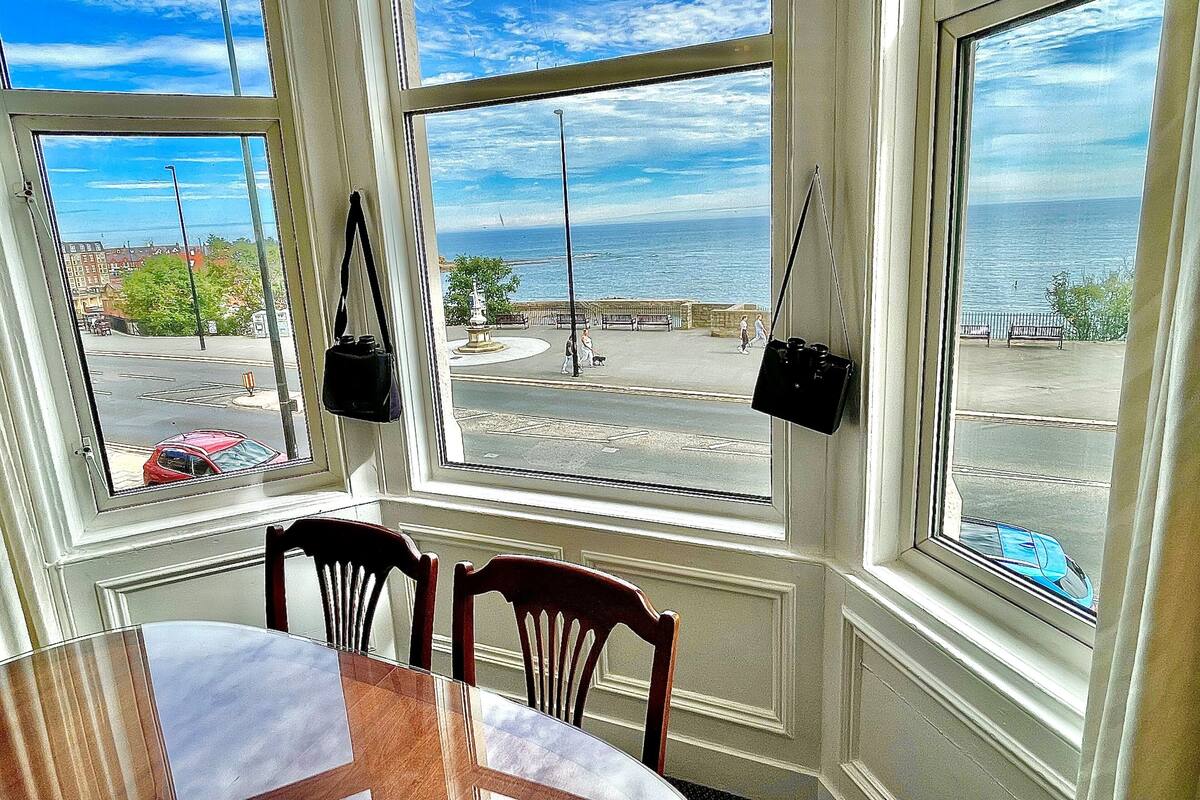 A room with a table and chairs, large windows with views of the street and sea, and a hanging bag.