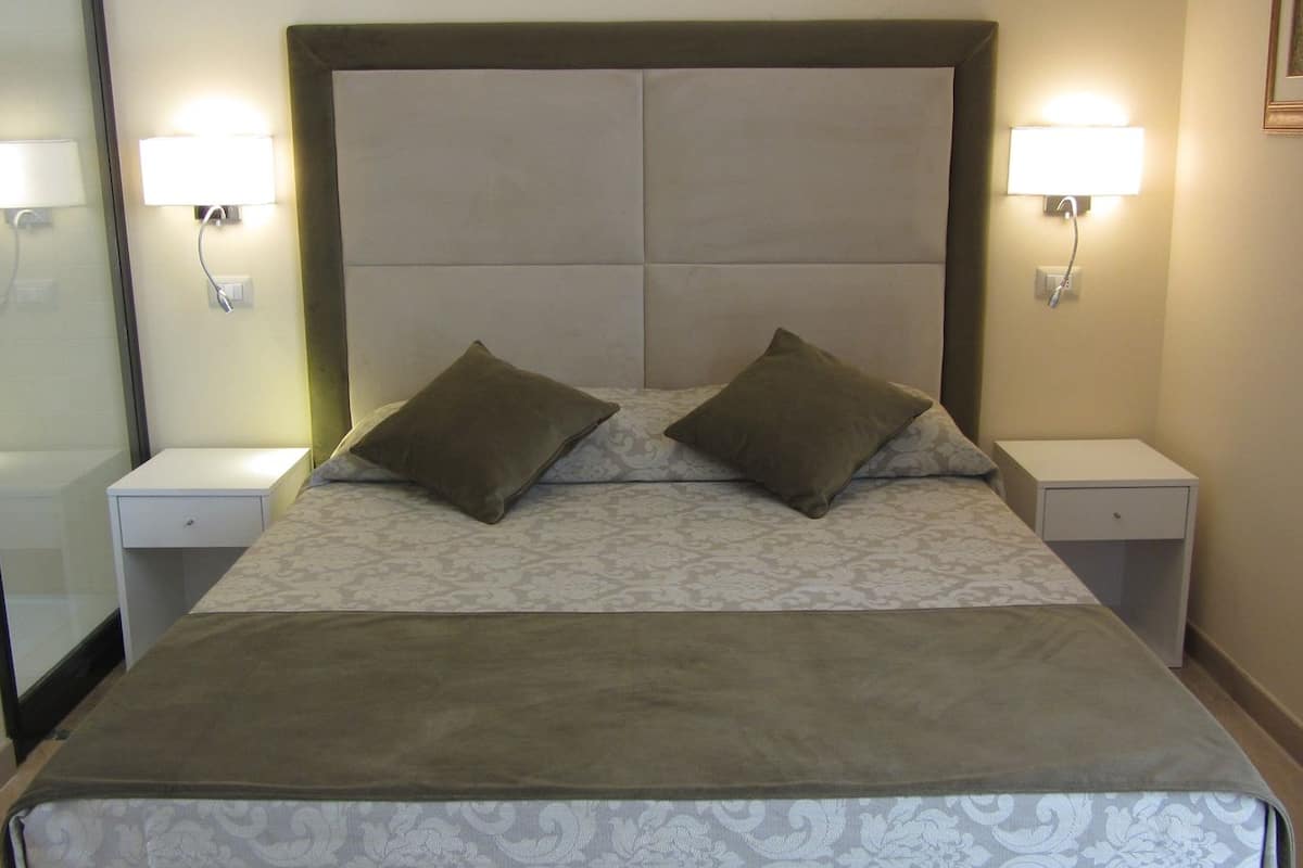 Una cama bien hecha con cabecera, dos lámparas de noche y una mesita de noche con cajón.