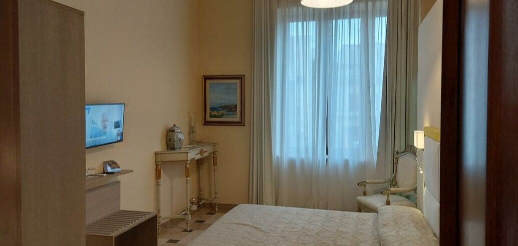 A hotel room with a bed, a desk with a lamp, a chair, a television, and a window with curtains.