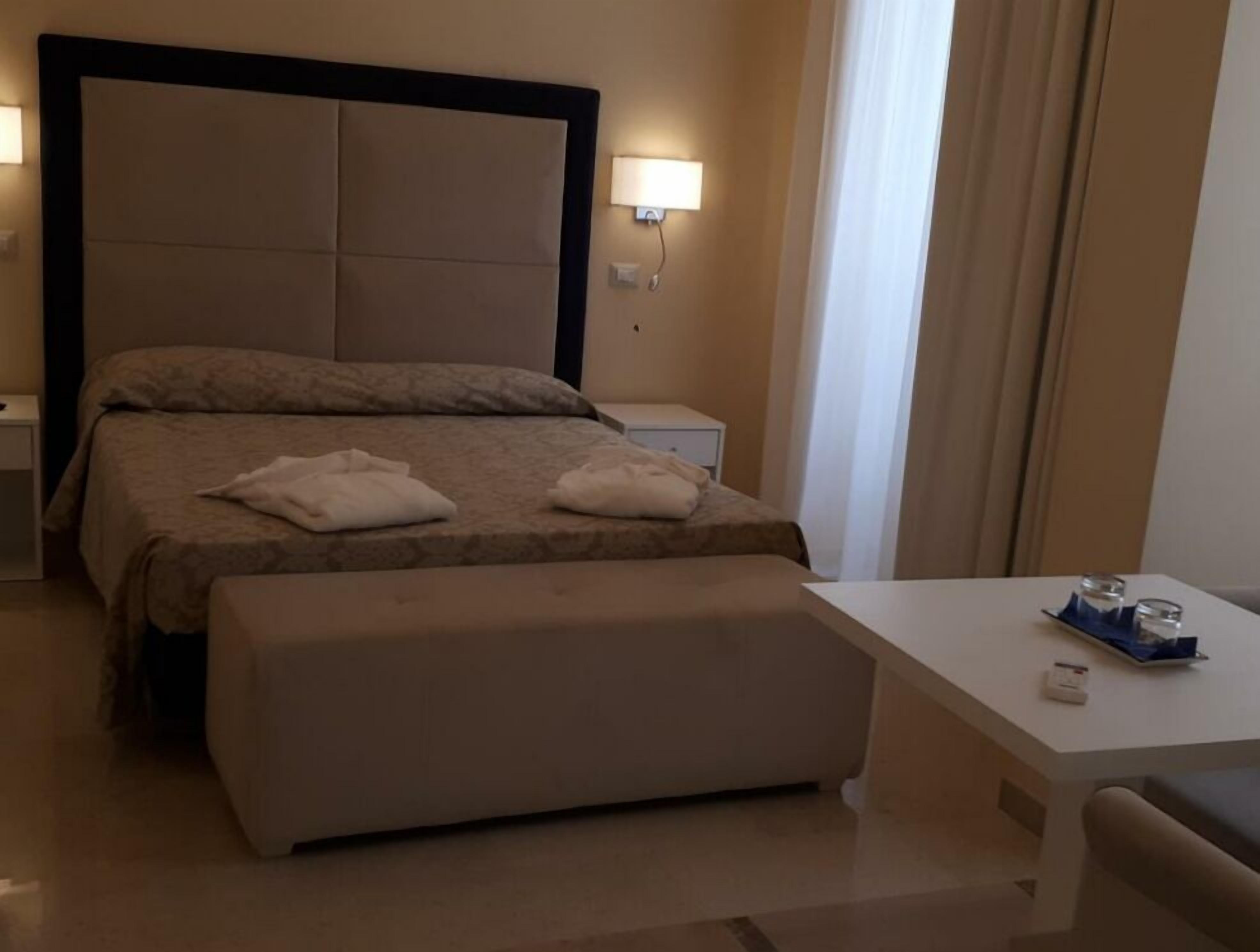 A hotel room with a large bed, a bedside table, a small table with a tray, and a window with curtains.