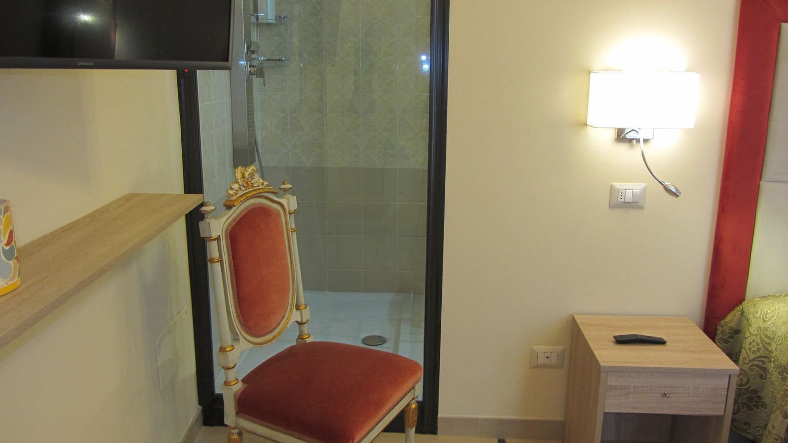 A bathroom with a shower, a chair, a bedside table, and a wall-mounted light.