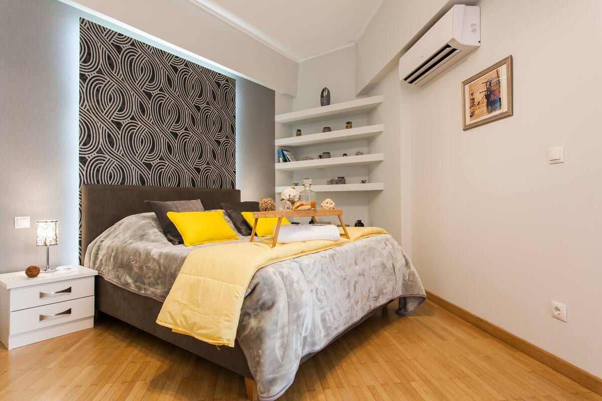 A modern bedroom with a grey bed, yellow and grey bedding, a bedside table with a lamp, and a patterned wall.