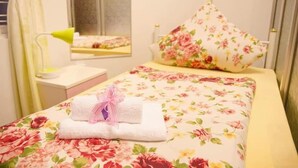 A hotel room with a floral bedspread, a neatly folded towel with a pink ribbon, a floral pillow, and a mirror above the bed. - Adler Lady Guesthouse - Women Only (Yangon)