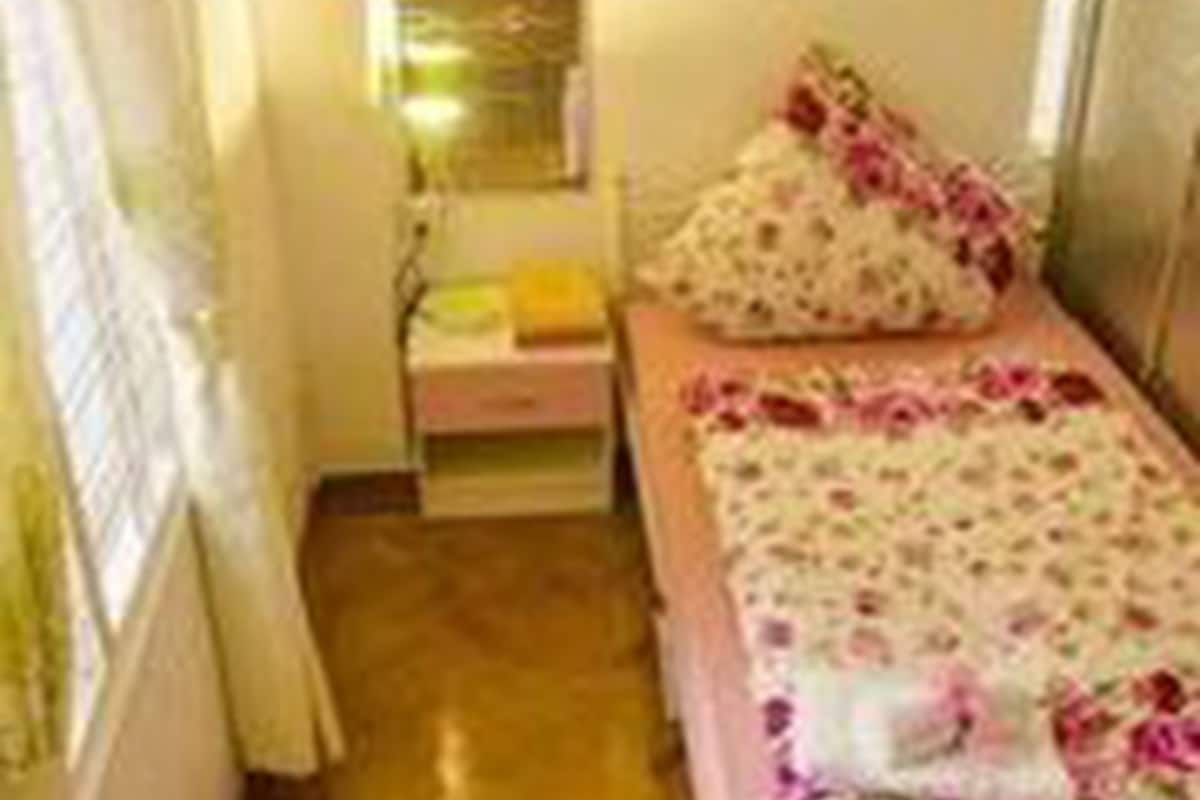 A small, narrow room with a single bed, a nightstand, a mirror, and a window with curtains.