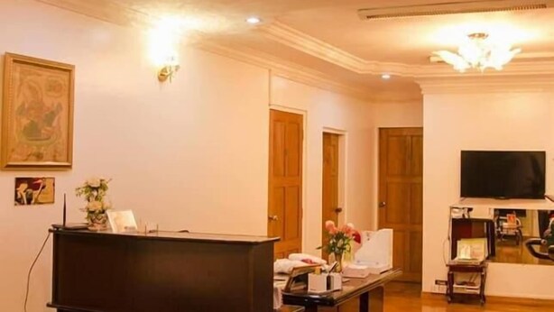 A spacious living room with a dark wooden reception desk, a mounted television, and a chandelier. - Adler Lady Guesthouse - Women Only (Yangon)