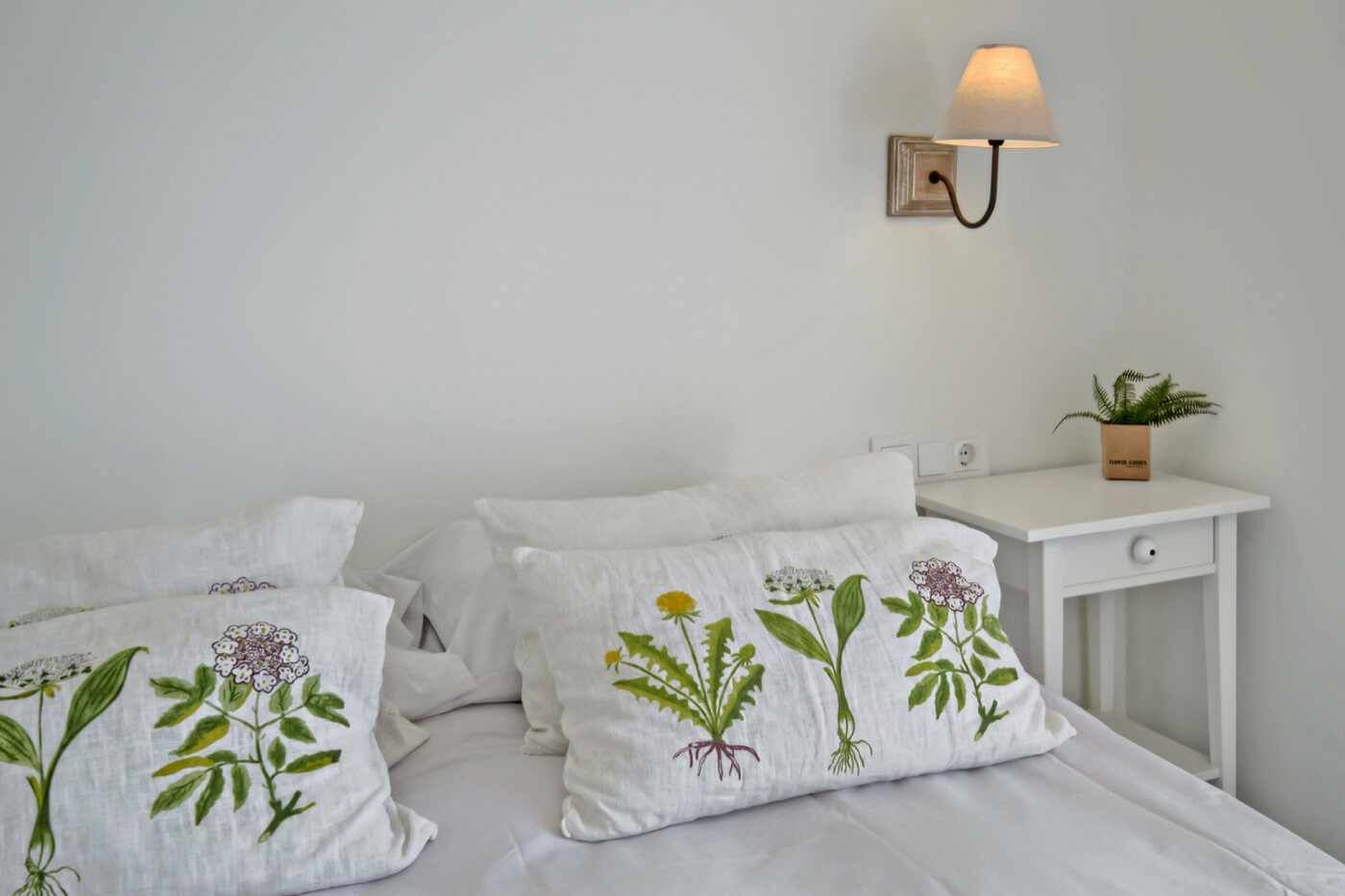 Un dormitorio con cama, almohadas de diseño floral, una mesita de noche con lámpara y una planta pequeña.