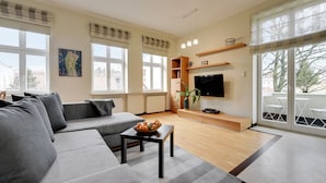 Una sala de estar moderna con un sofá gris, una mesa de centro de madera con fruta, un televisor de pantalla plana y ventanas grandes.
