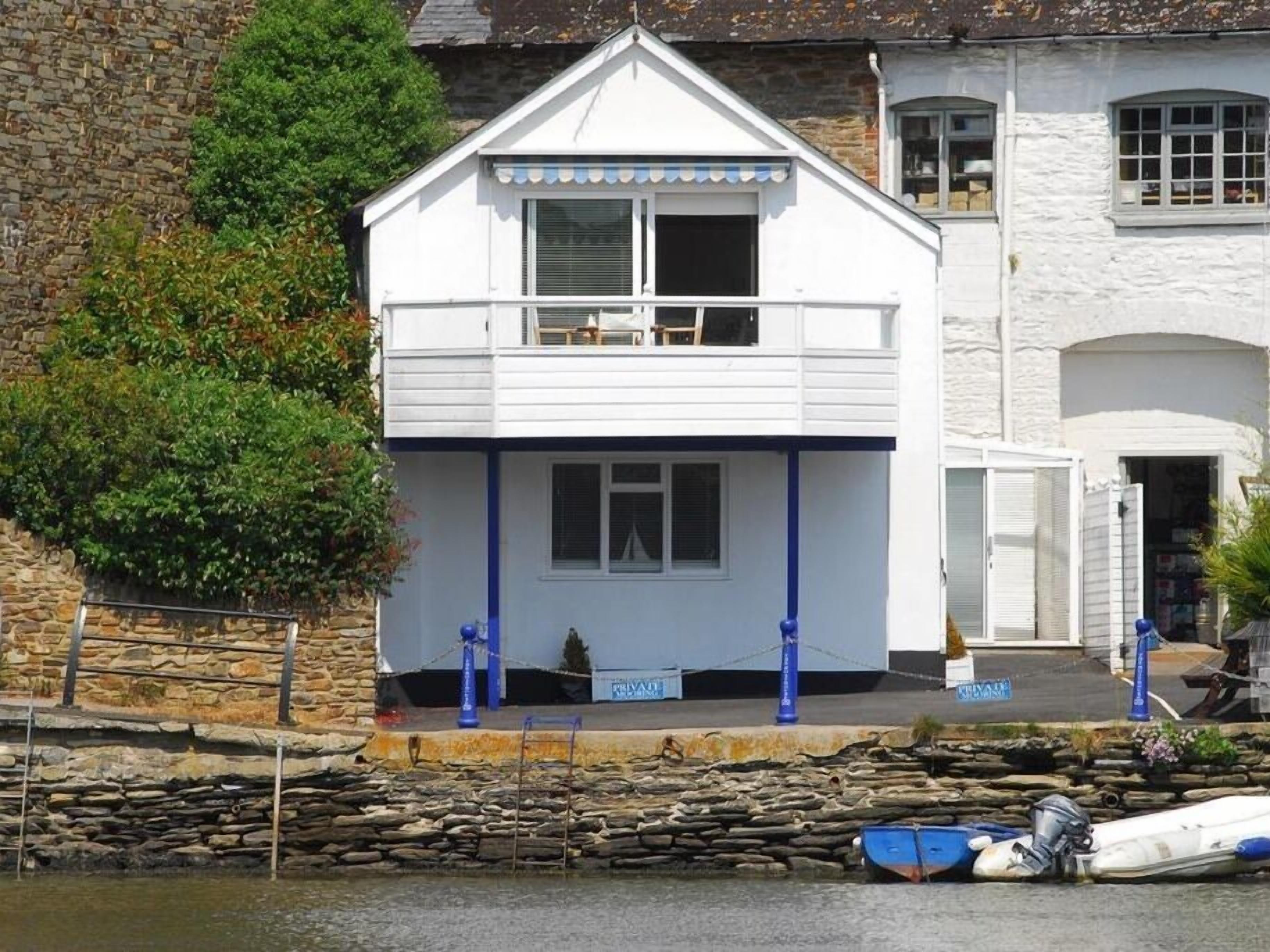 Un bâtiment blanc doté d’un balcon, d’un mur en pierre et d’un petit bateau amarré devant.