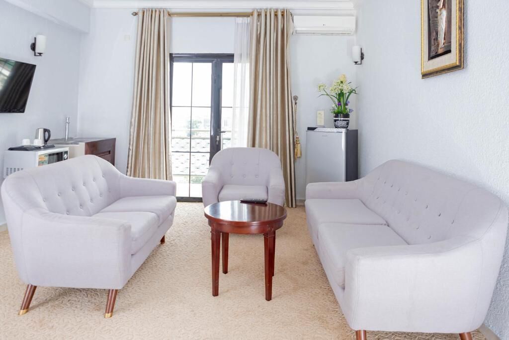 Habitación de hotel con dos sofás blancos, una mesita de madera, televisor, minibar y vista a la ciudad desde la ventana.