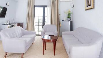 Habitación de hotel con dos sofás blancos, una mesita de madera, televisor, minibar y vista a la ciudad desde la ventana.