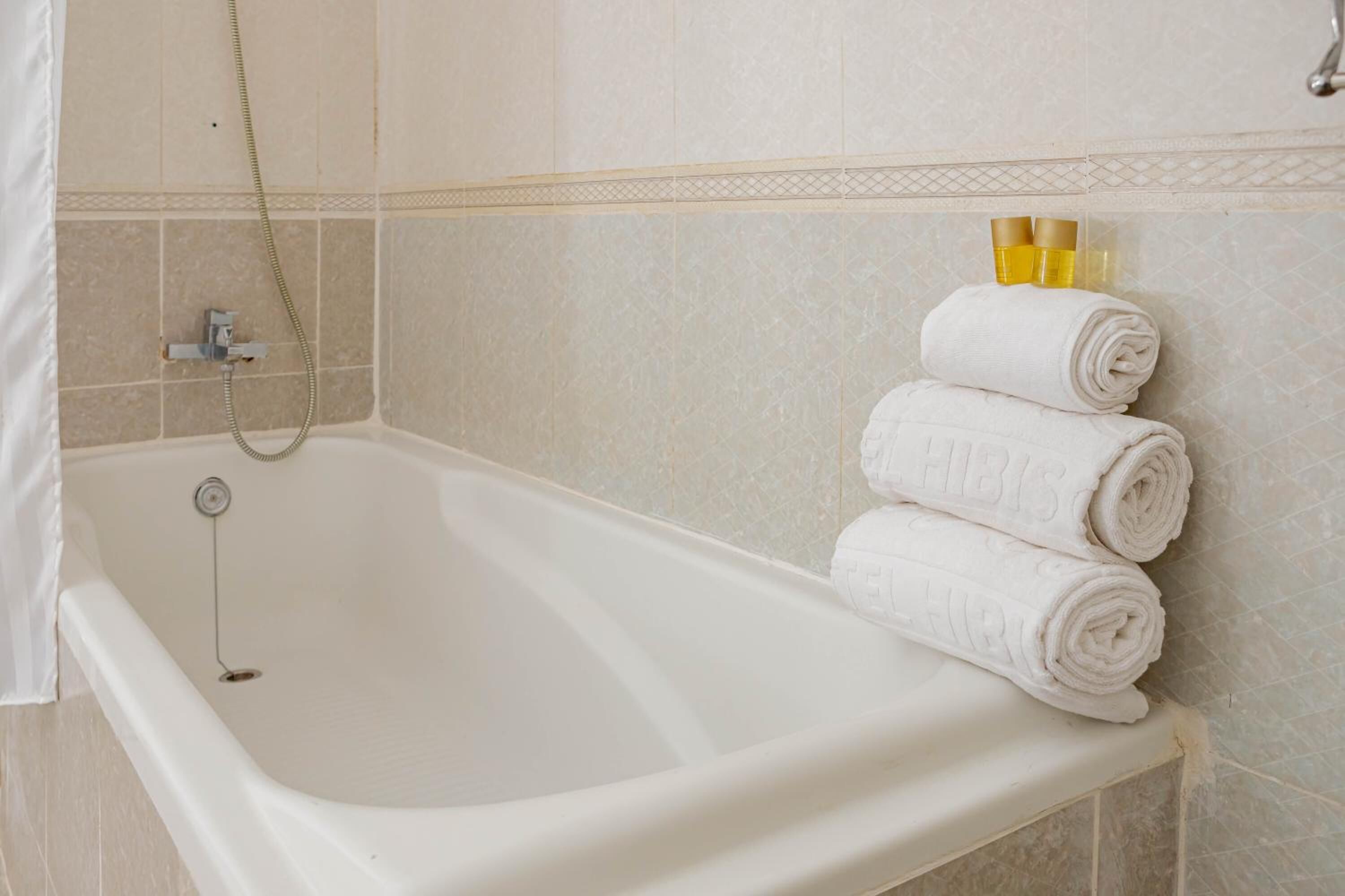 A white bathtub with a showerhead, two rolled towels, and two bottles of lotion on a shelf.