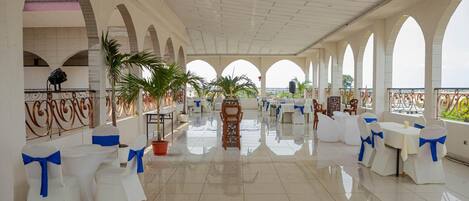 A spacious indoor area with white tables and chairs, some with blue sashes, and a tiled floor.