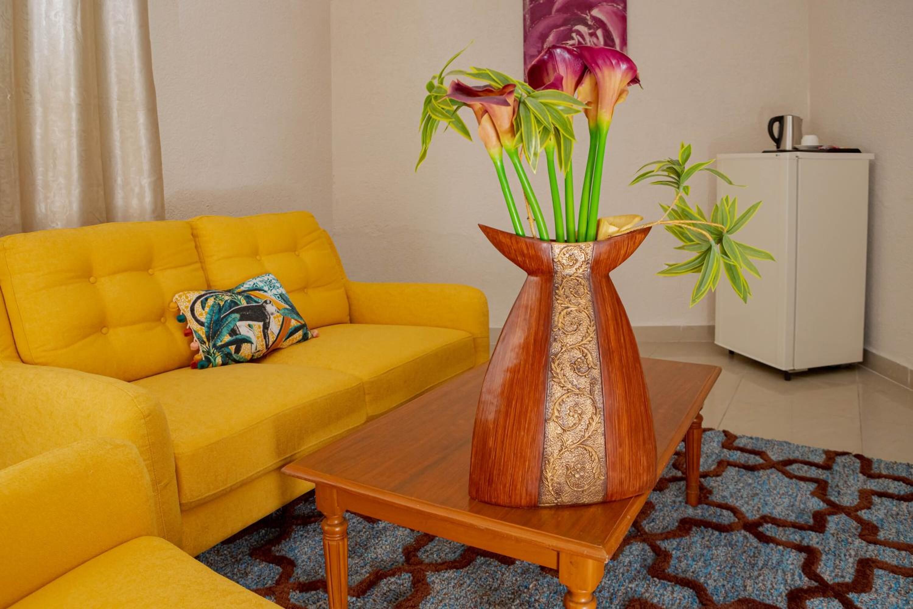 A yellow sofa with a patterned pillow, a wooden coffee table, a vase with flowers, and a white cabinet.
