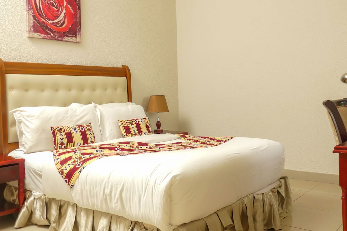 A neatly made bed with white linens and patterned pillows, a wooden headboard, a bedside lamp, and a red abstract painting on the wall.