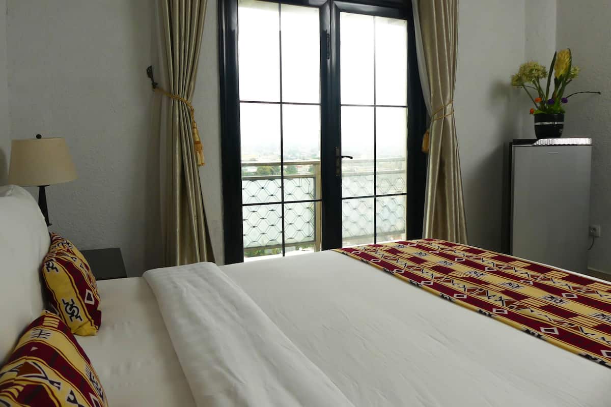 A hotel room with a bed, curtains, a window with a view, a lamp, and a refrigerator with flowers.
