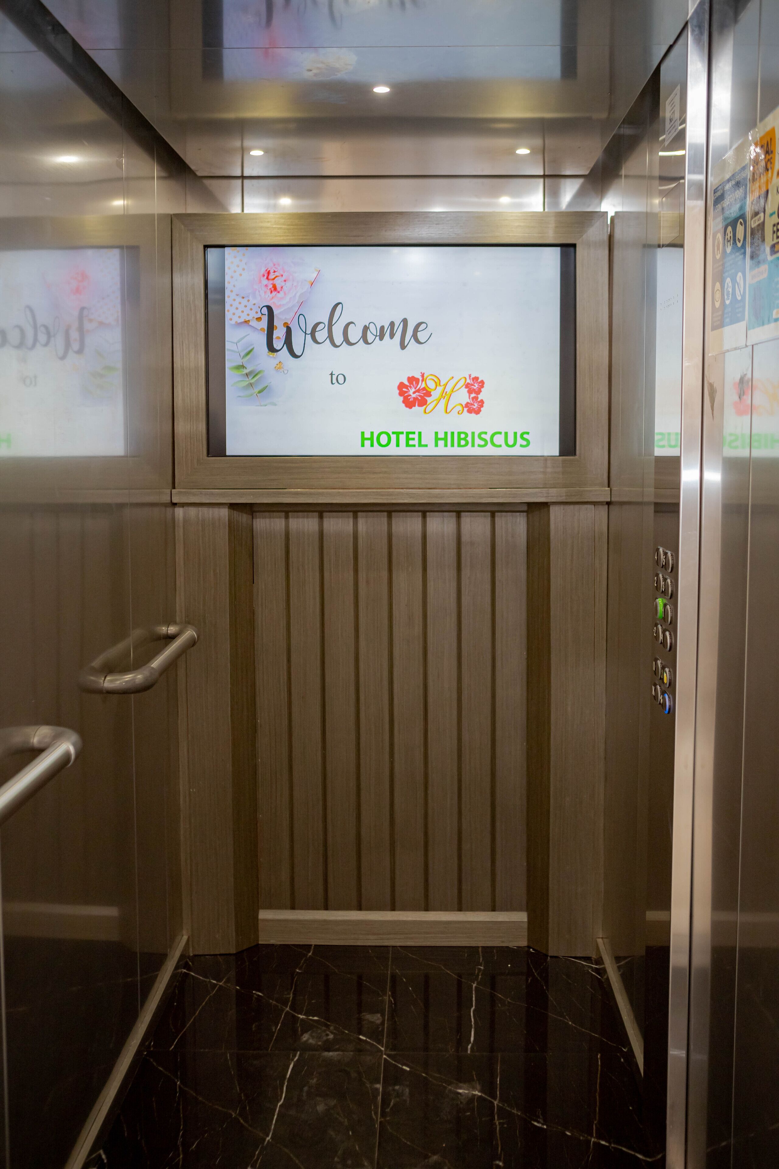 An elevator with a welcome sign for HOTEL HIBISCUS, featuring a modern design with a digital display and metallic interior.