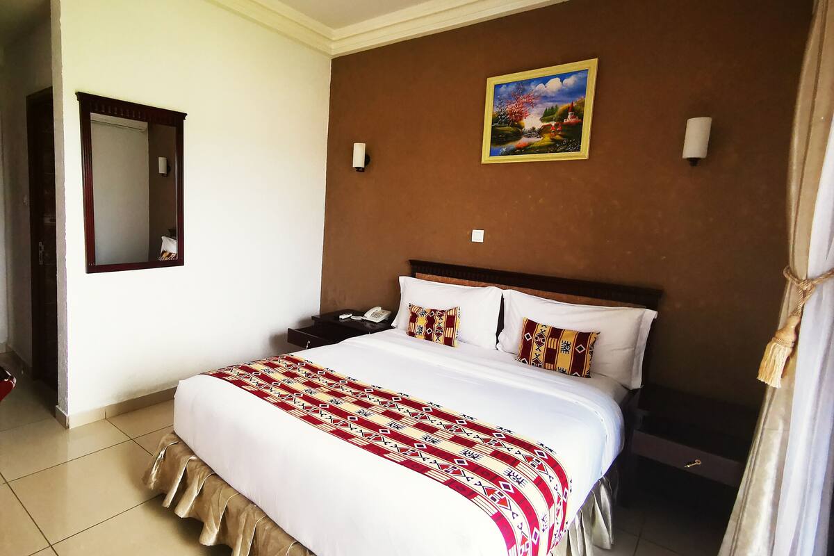 A hotel room with a large bed, a nightstand with a phone, a wall-mounted lamp, and a painting above the bed.