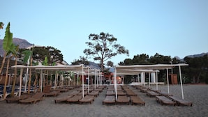 A beach with wooden lounge chairs and white canopies, a mountain in the background, and a tree in the center. - Monna Roza Beach Resort Hotel (Kemer)