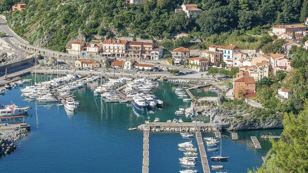 Un pueblo costero con un puerto deportivo lleno de barcos, rodeado de edificios residenciales y un paisaje montañoso.