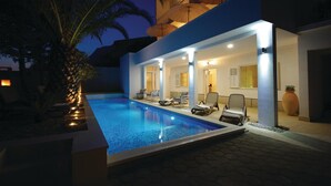 A modern poolside area with lounge chairs and umbrellas, a well-lit pool, and a building with a balcony.