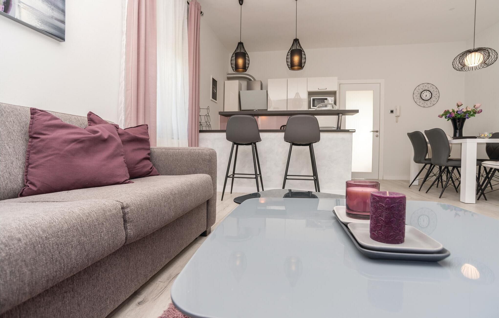 A modern living room with a grey sofa, two black bar stools, a glass coffee table with candles, and a kitchen in the background.