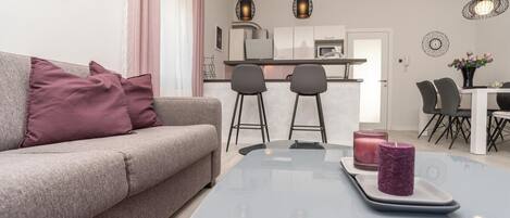 A modern living room with a grey sofa, two black bar stools, a glass coffee table with candles, and a kitchen in the background.
