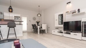 Une cuisine moderne avec des meubles blancs, un coin repas avec une table et des chaises blanches, un téléviseur posé sur un meuble blanc et un bar avec des tabourets.