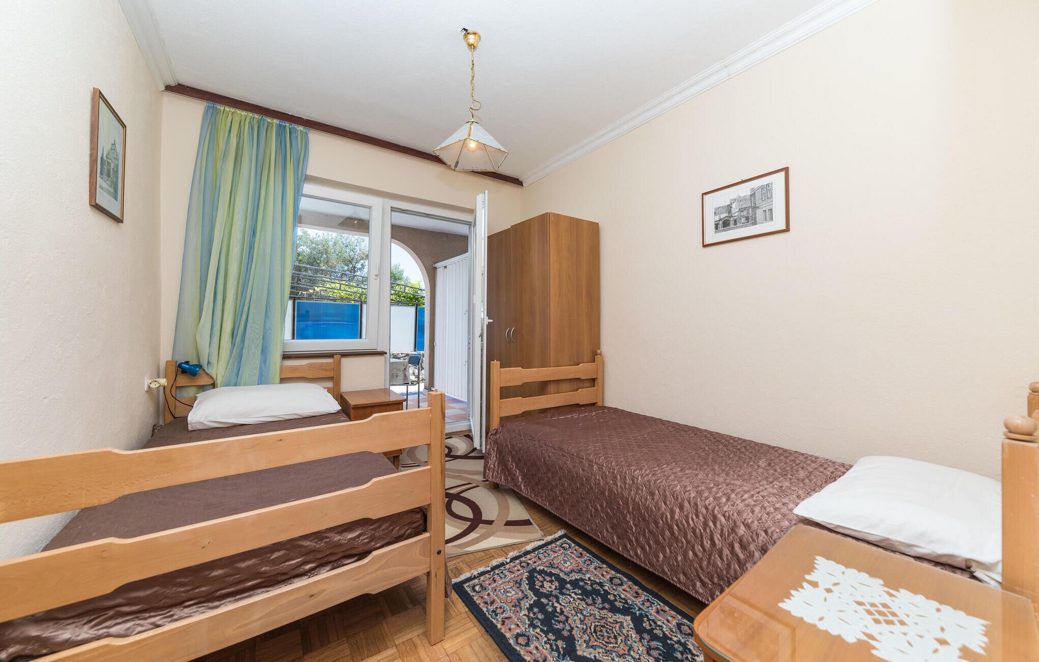 Una habitación pequeña y ordenada con dos literas de madera, una cama individual, un armario de madera, una lámpara en el techo y una ventana con cortinas.
