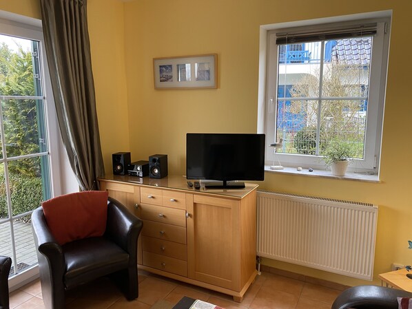 A room with a TV on a wooden cabinet, a chair, a window with a view of trees, and a radiator. - Bright, family-friendly apartment near the beach (Dassow)