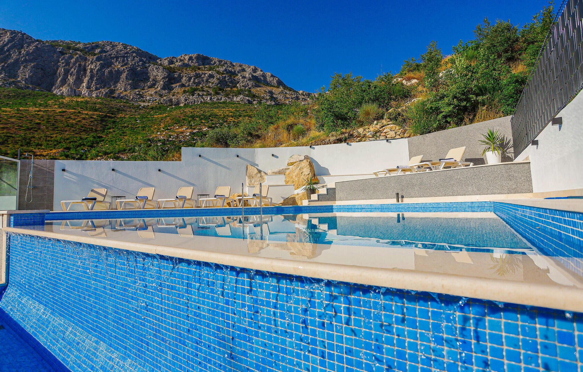 A swimming pool with clear blue tiles, surrounded by lounge chairs and a view of rocky mountains.