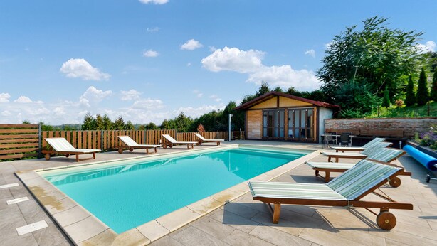 Une piscine avec des chaises longues et un petit bâtiment, le tout entouré d’une clôture en bois et de verdure.