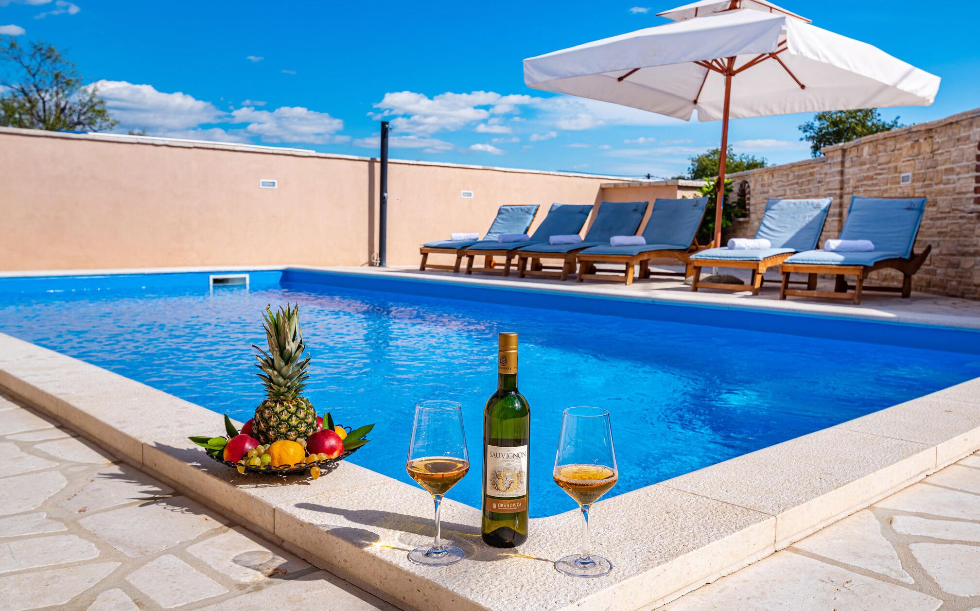 A swimming pool with lounge chairs and umbrellas, a bottle of wine, and a fruit basket.