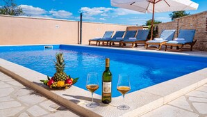 A swimming pool with lounge chairs and umbrellas, a bottle of wine, and a fruit basket. - Amazing home in Pridraga (Pridraga)